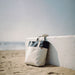 Field & Co.® Parks Day Tote - A beach tote bag in cream and navy rests against a white wall on sandy shore, with the ocean in the background.