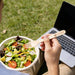 Pack of 100 Per Pack Disposable Wooden Cutlery - (Forks /Spoons/Knives/2 in 1 Spoon and fork)-(Big) - A person holding a wooden fork over a salad bowl, with a laptop in the background on grass. The fork has a logo.