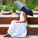 Poppins Umbrella - A black umbrella rests over a person wearing a white dress, seated on wooden decking surrounded by planters.