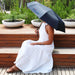 Poppins Umbrella - A black umbrella with a logo, held by a person in a white dress, sitting on wooden steps with greenery in the background.
