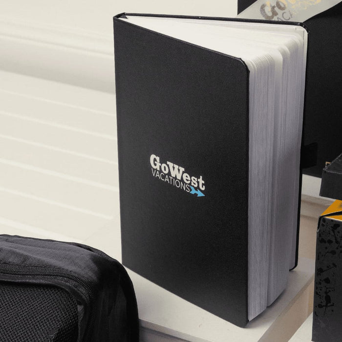 The Custom Travel Kit - A black notebook with a logo, featuring blank pages, rests on a white surface alongside other items.