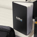 The Custom Travel Kit - A black notebook with a logo, featuring blank pages, rests on a white surface alongside other items.