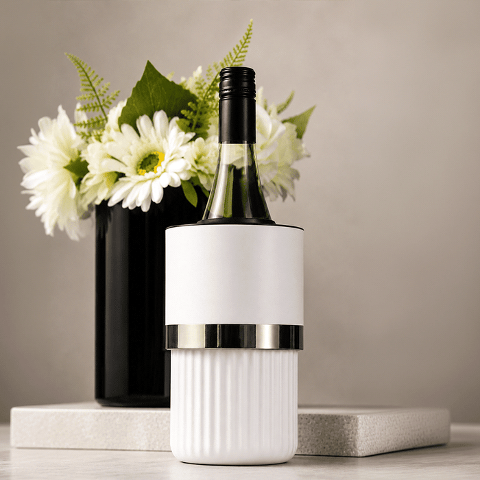 Trekk Insulated Adjustable Wine Cooler - A white wine bottle cooler with a silver band, placed on a marble surface beside a black vase with white flowers.