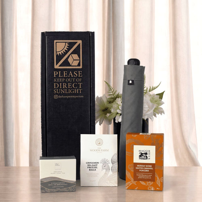 The Blunt Umbrella All Weather Set - A grey umbrella stands beside packaged tea and coffee products, all displayed in a black box, with flowers in the background.