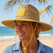 Beach Straw Hat - A straw beach hat with a yellow brim and chin strap, worn by a woman at the beach with palm trees in the background.