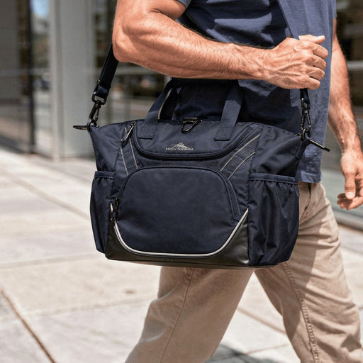 High Sierra Crib Bag - A navy cooler bag with a sturdy strap is being carried by a person walking on a city sidewalk. It has a logo.