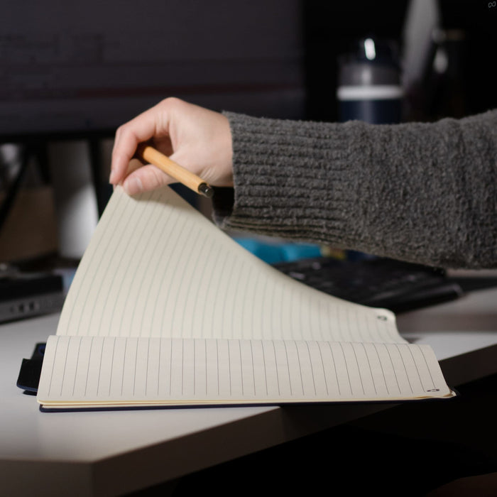 SunScribe Notebook (A4) - A hand holding a pencil opens a lined A4 notebook with a dark blue cover on a desk.