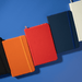 Minichroma A6 Notebook - A collection of Minichroma A6 Notebooks in black, orange, red, navy, and white, arranged on a blue surface.