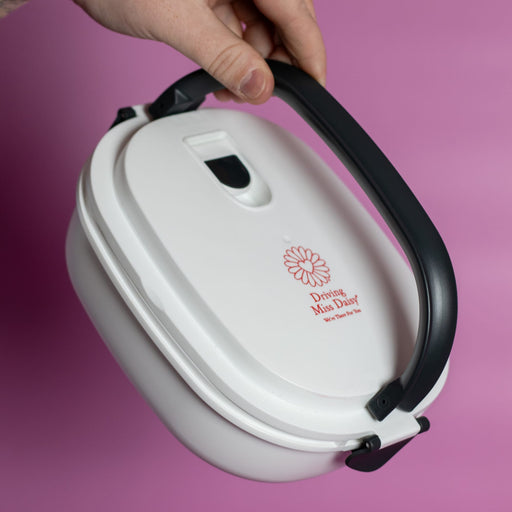 The Lunch Box 800ml - A white lunch box with a black handle, featuring a floral logo, held against a pink background.