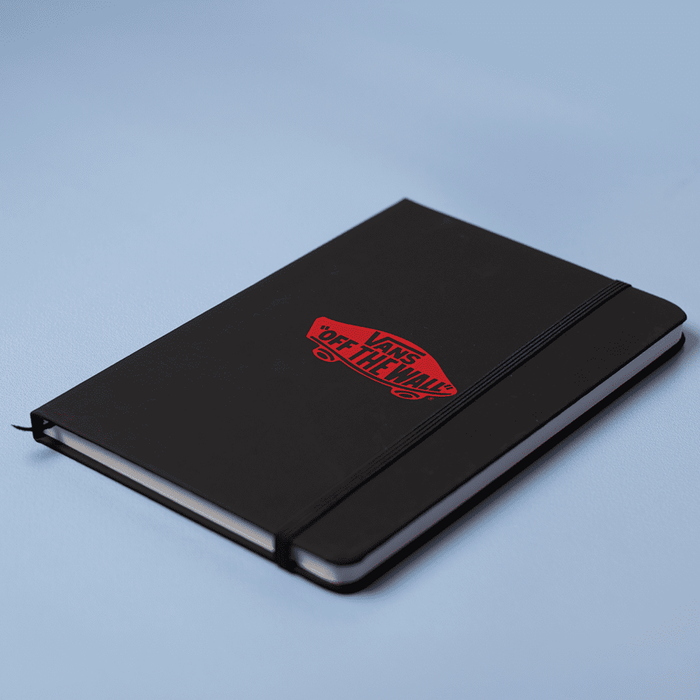 Space A5 Notebook - A black A5 notebook with a logo on the cover and an elastic closure, set against a light blue background.