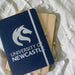 Discovery A5 Notebook - A navy A5 notebook with a logo, placed atop a beige notebook, surrounded by flower petals on a soft surface.