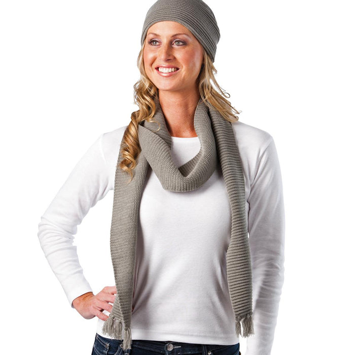 Ruga Knit Scarf - A woman wearing a grey knitted scarf and beanie, with her hair cascading down and a white long-sleeve top.
