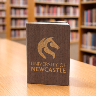 The Barker Eco Notebook - B6 - A brown eco notebook with a logo on the cover, placed on a wooden table in a library setting.