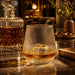 Velora Tumbler by The Line Edit. - A drink glass with a ridged texture, filled with amber liquid, sits on a marble surface beside a crystal decanter.