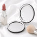 Finesse Compact Mirror - A compact mirror with a sleek silver frame, accompanied by a lipstick and a makeup brush on a soft white fabric background.