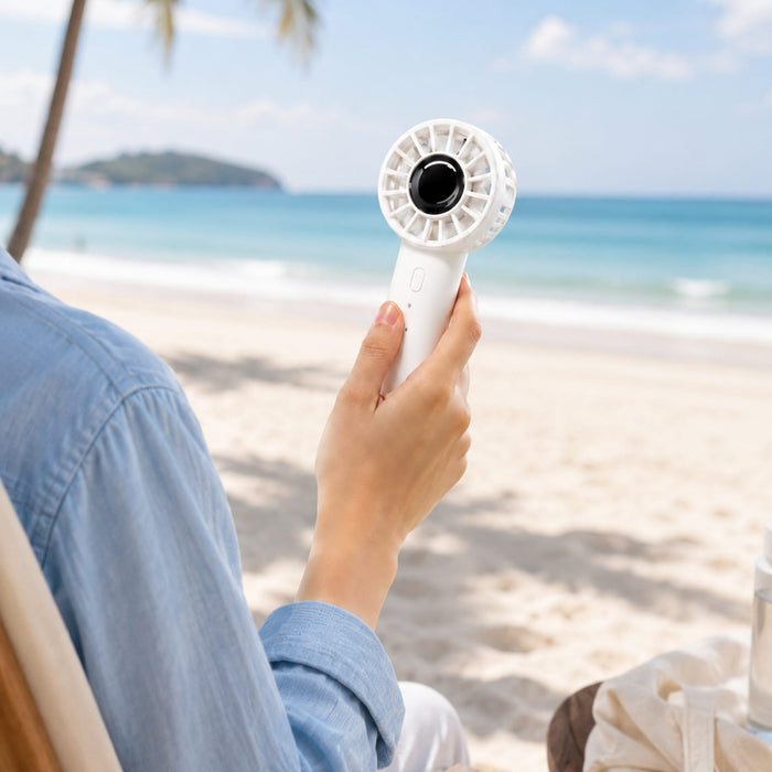 Nomad Rechargeable Fan - A white rechargeable fan is held by a person, set against a beach background with sand and palm trees.