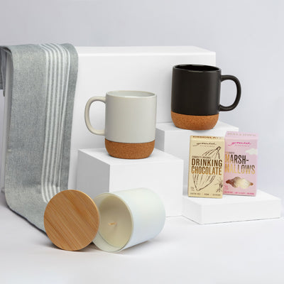 The Warm Welcome - A collection of two mugs in grey and black with cork bases, a candle, and chocolate and marshmallow packets on white display.