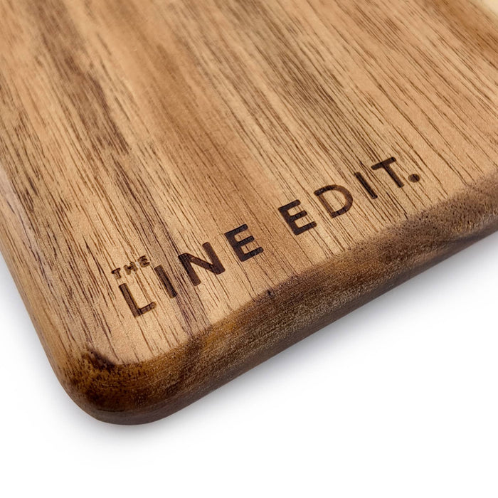 The Line Edit Acacia Serving Board - A wooden serving board with a smooth acacia finish featuring a branding logo.