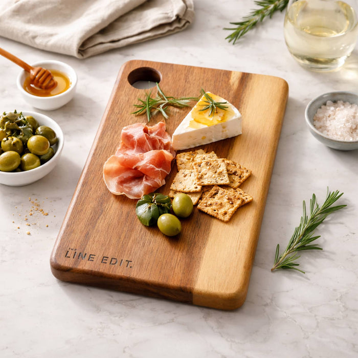 The Line Edit Acacia Serving Board - A wooden serving board featuring cured meats, cheese, crackers, olives, and fresh herbs, with a minimalist branding logo.