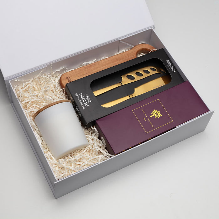 The Host Edit - A gift box containing a cheese knife set, a wooden serving board, and a cup, arranged with white filler.