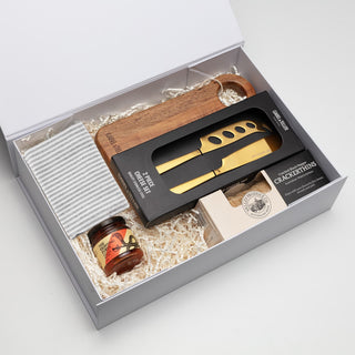 The Nibble Set - A gift box containing a wooden cheese board, a gold cheese knife, honey, and crackers, arranged on straw.