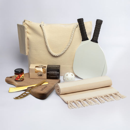 The Sunchaser Set - A cream tote bag, wooden board, drink jar, cheese knife, paddles, ball, picnic blanket, and gourmet snacks.