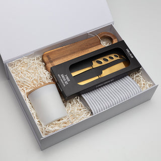 Glow & Graze - A wooden cheese board, a gold knife, a white cup, and a grey towel in a gift box with straw filling.
