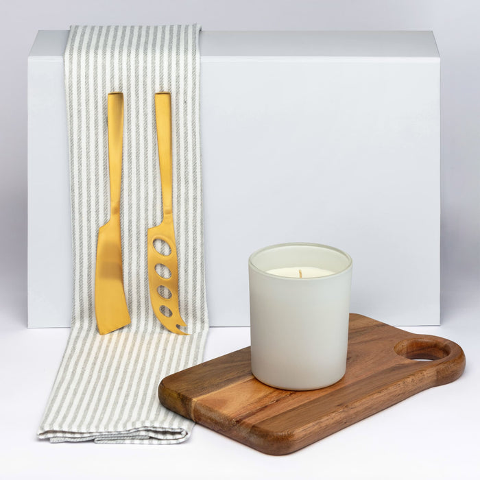 Glow & Graze - A wooden serving board, a white candle in a frosted glass holder, and golden utensils on a striped cloth.