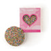 The Generous Host - A round cookie covered in colourful milk chocolate sprinkles next to a pink box with a heart-shaped window.