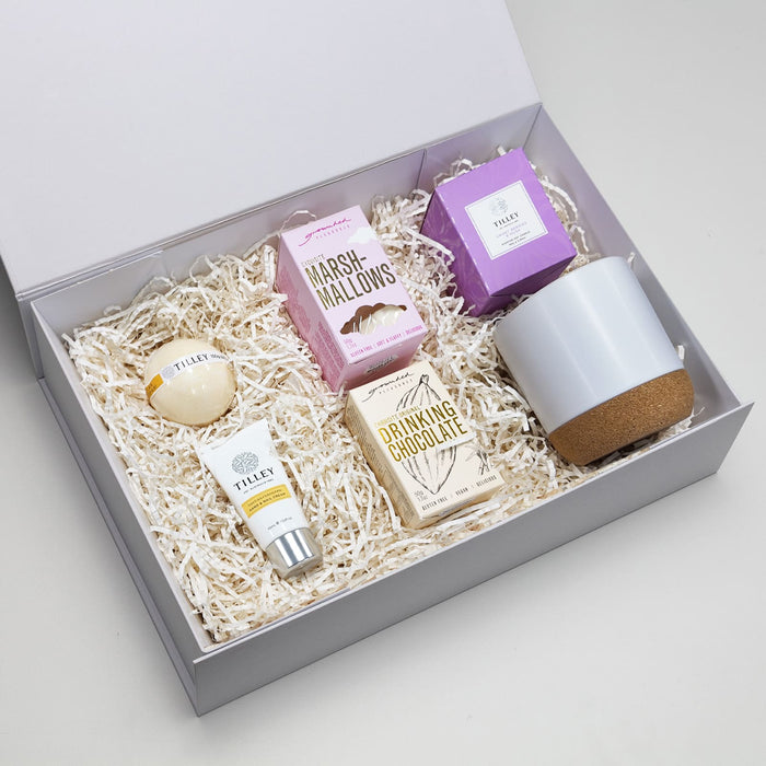 The Cosy Indulgence - A gift box containing a grey pottery cup, drinking chocolate, marshmallows, and assorted skincare items.