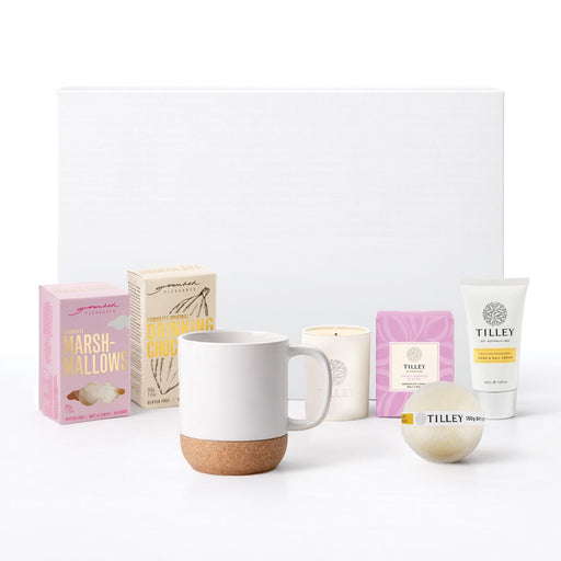 The Cosy Indulgence - A collection of skincare products, a candle, a mug with a cork base, and decorative boxes in pastel colours.