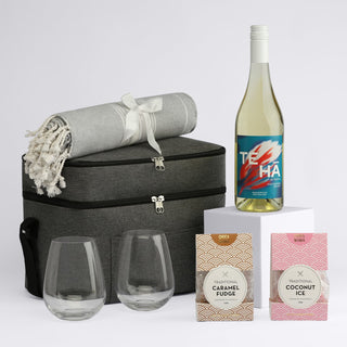 The Wine & Unwind - A drink bottle and two stemless glasses sit beside a picnic basket, blanket, and two gourmet treats in pink packaging.