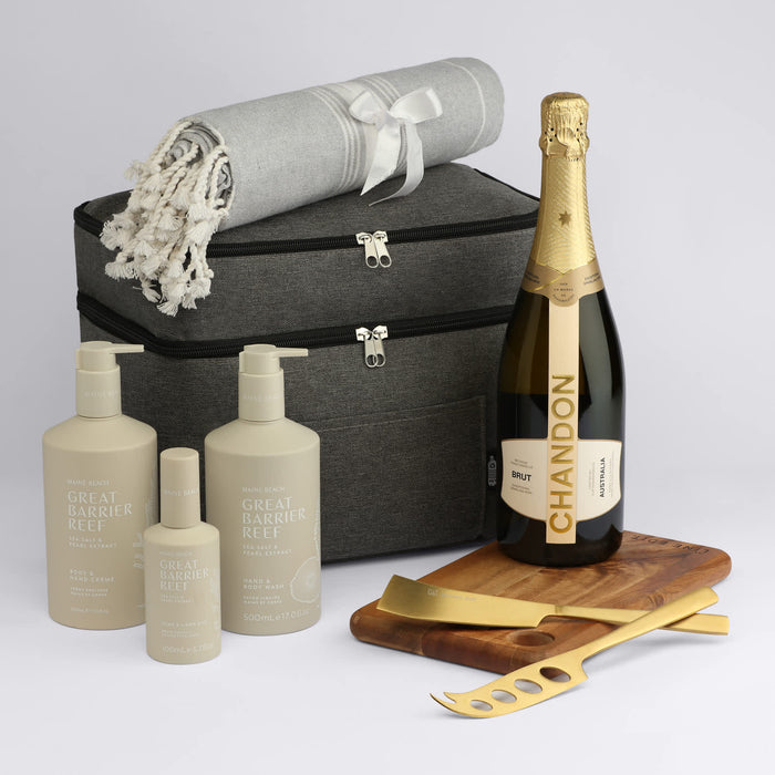 The Coastal Edit - A grey storage bag contains a throw blanket, champagne bottle, and skincare products in elegant packaging on a wooden tray with serving tools.