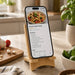 Oscar Illuminating Charging Stand - A wooden charging stand with a smartphone displaying a pasta recipe, surrounded by kitchen items and greenery.