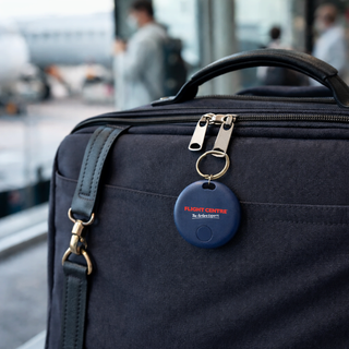 SeekTag Factory Direct - A navy blue tracking device attached to a dark bag, set against a blurred airport background.