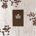 Coffee Grounds A6 Notebook - A brown A6 notebook surrounded by coffee beans on a wooden surface, featuring a logo on the cover.