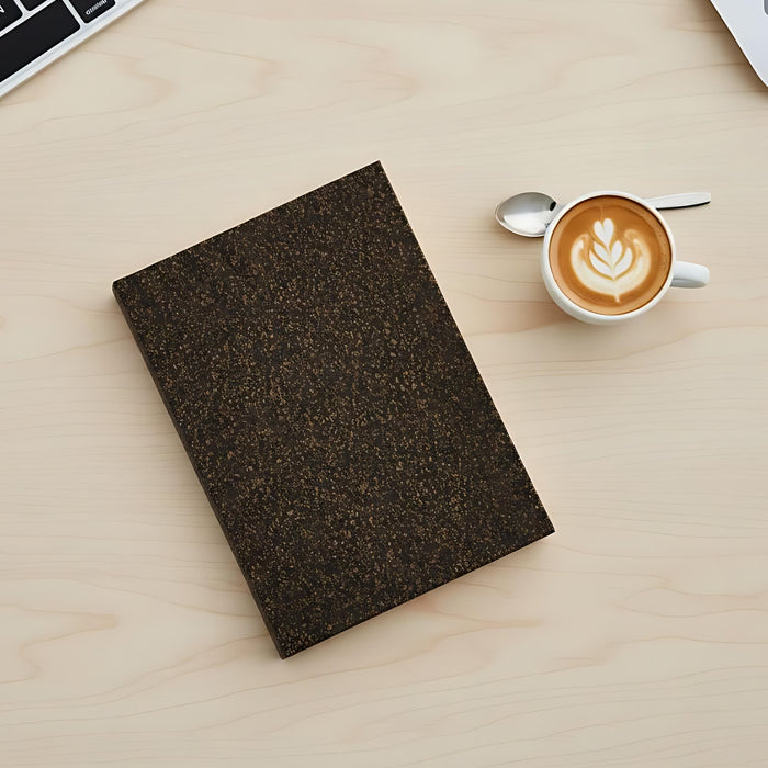 Coffee Grounds A6 Notebook - A dark brown notebook with a textured cover rests on a light wooden surface next to a cup of latte art.