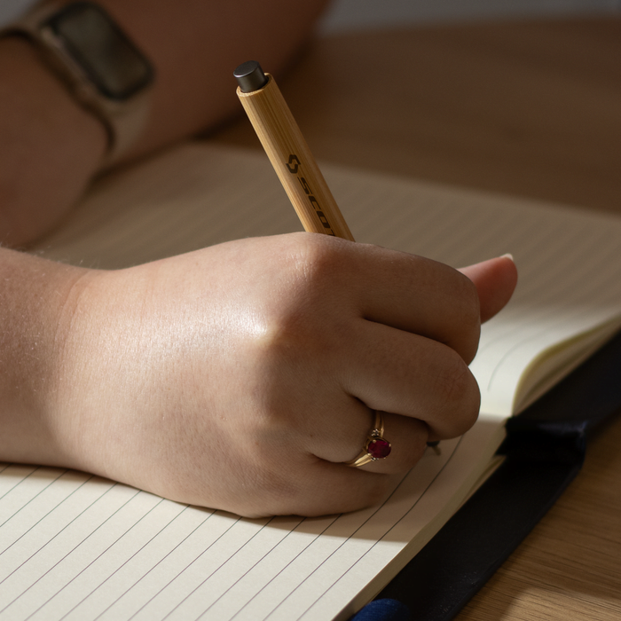 SunScribe Notebook (A4) - A hand holding a pen writes in a lined, cream-coloured notebook on a wooden surface.