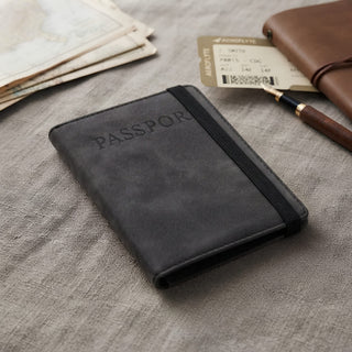 Traveller Passport Wallet - A black passport wallet with an elastic band, resting on a textured surface alongside travel documents and a pen.