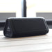 Trekk Outdoor Speaker & Wireless Powerbank - A black outdoor speaker with a triangular shape and a logo, resting on a wooden surface.