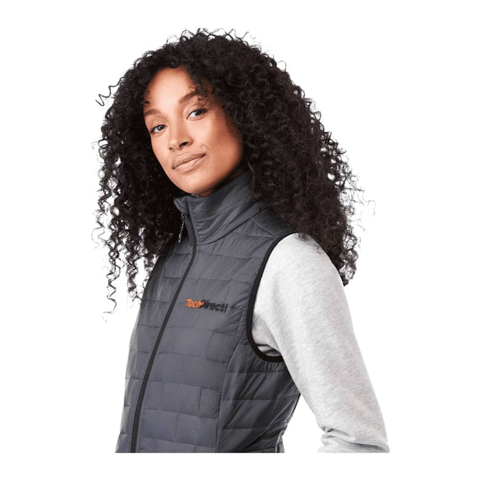 Women's Telluride Lightweight Packable Insulated Puffer Vest - A woman with curly hair wears a black insulated puffer vest, featuring a logo on the chest.