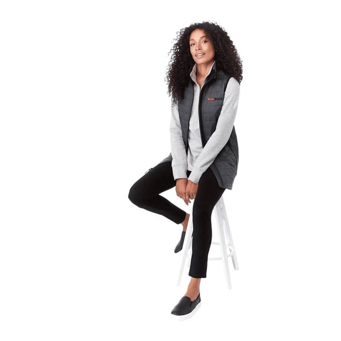 Women's Telluride Lightweight Packable Insulated Puffer Vest - A woman sits on a stool wearing a black insulated puffer vest over a light grey top, paired with black trousers.