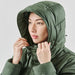 Women's Elkridge Parka - A woman wearing a green puffer jacket with a hood, adjusting it around her face. The jacket has a subtle logo.