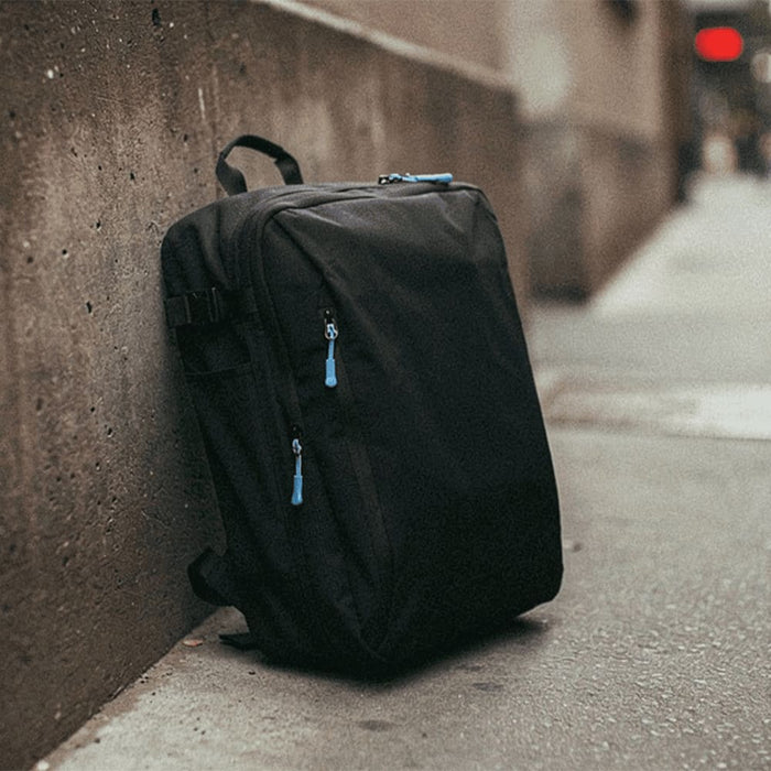 Wander & Roam Travel Backpack - A black travel backpack with blue zipper pulls leaning against a concrete wall. It features multiple compartments.