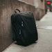 Wander & Roam Travel Backpack - A black travel backpack with blue zipper pulls leaning against a concrete wall. It features multiple compartments.